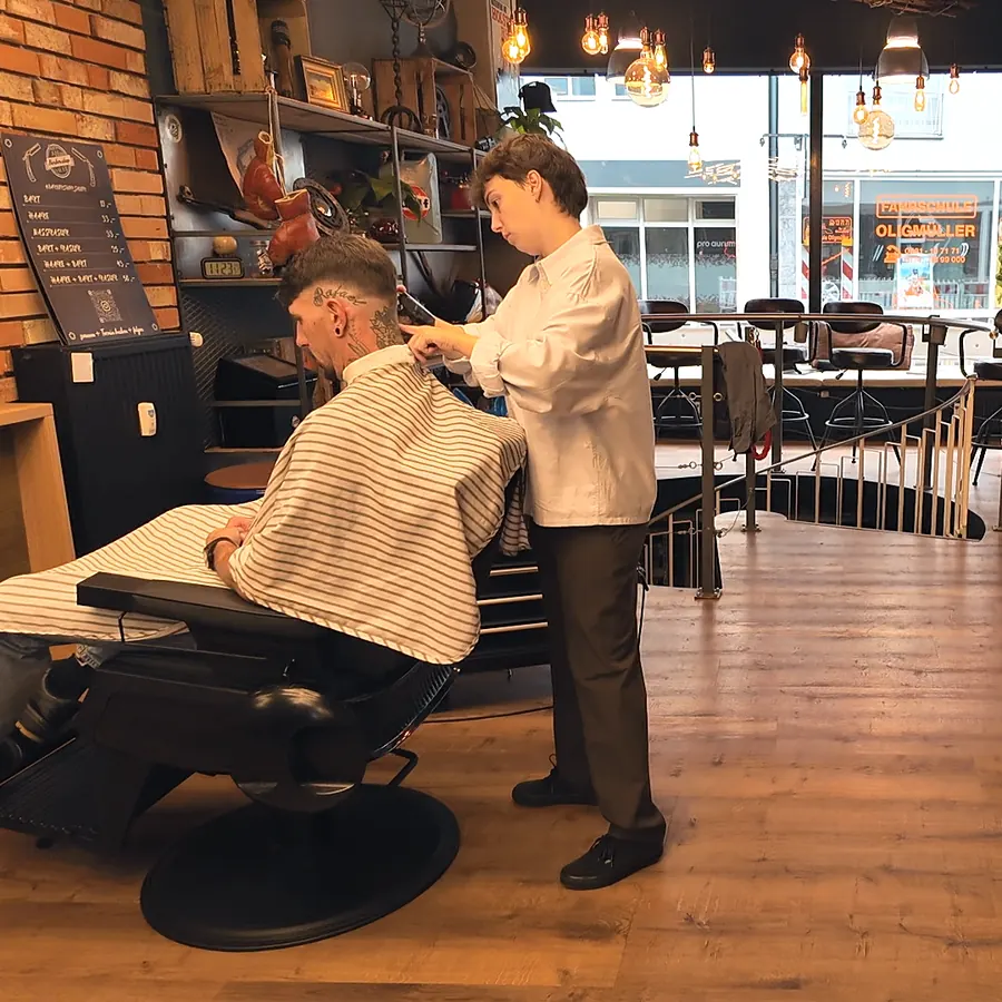 Barber im weißen Hemd schneidet einem Kunden im Barberstuhl die Haare, dahinter Ziegelwand, Regale und das Schaufenster