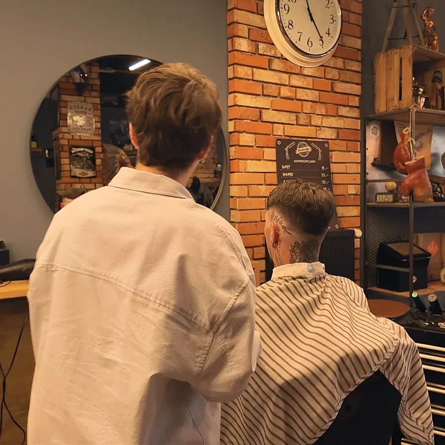 Barber im hellen Hemd von hinten bei der Arbeit, vor ihm ein Kunde im gestreiften Umhang, daneben Ziegelwand mit Uhr und runder Spiegel