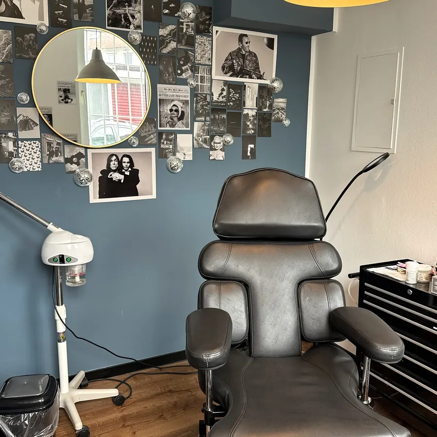 Raum für Men's Care: schwarzer Behandlungsstuhl, Gesichtsdampfer und runder Spiegel vor einer blauen Wand mit Schwarz-Weiß-Fotos