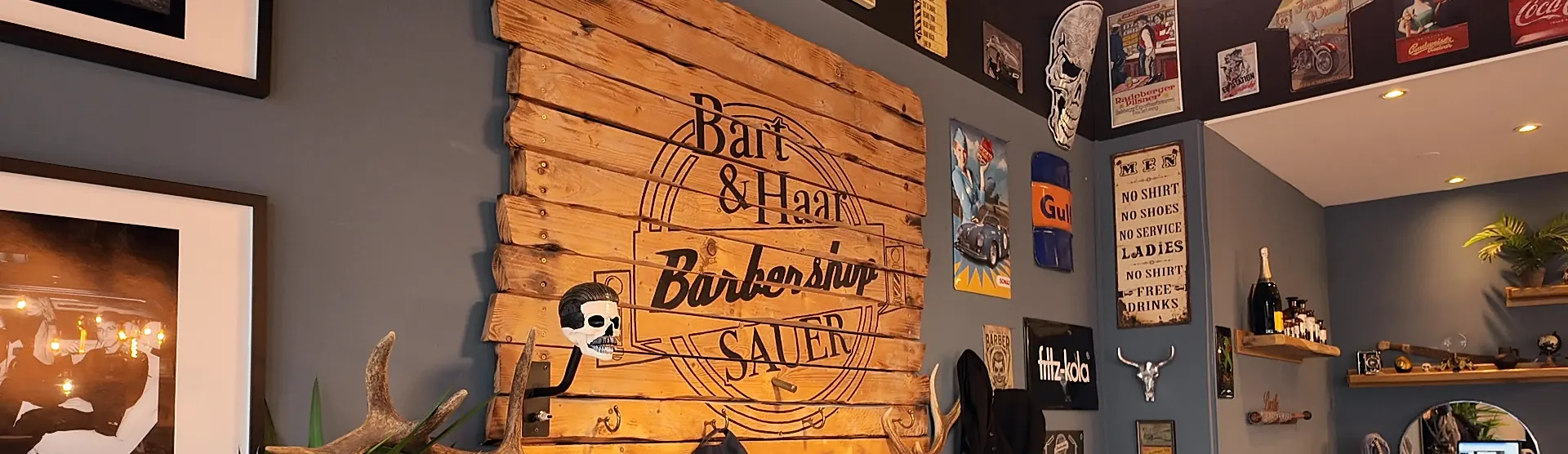 Holzschild an der Wand mit dem Schriftzug „Bart & Haar Barbershop Sauer“, daneben Blechschilder und Bilder